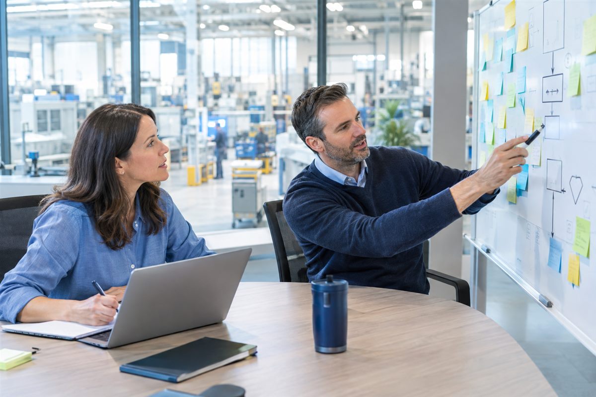 Quality consultant mapping a process on a whiteboard with a manager, factory floor in the background